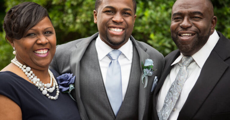 Groom and his parents standing close to each other with wide smiles showing their teeth.