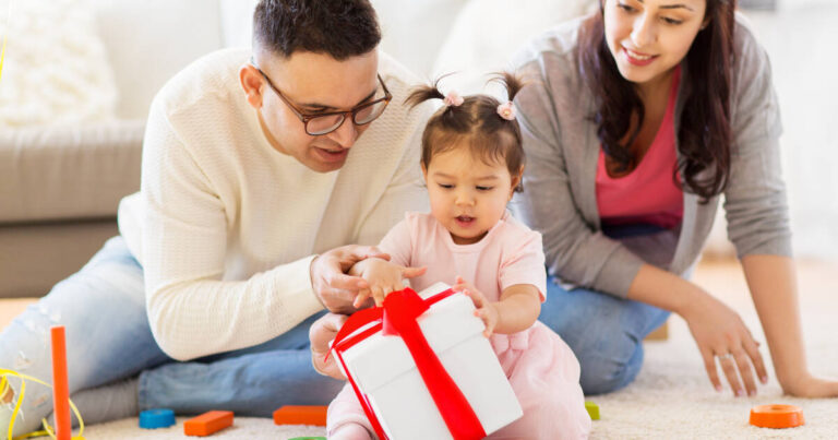 A young couple sits on the floor with their baby girl, helping her open a white gift box with a red ribbon. Colorful toys are scattered around them in a cozy home setting.