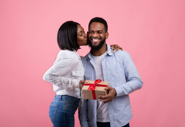 Woman kisses husband on cheek as he smiles, holding gift she just gave him on pink background