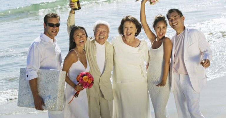 A joyful group stands on a sunny beach, dressed in white and celebrating a wedding. One holds a gift, another a champagne bottle, and the bride holds a bouquet with ocean waves in the background.