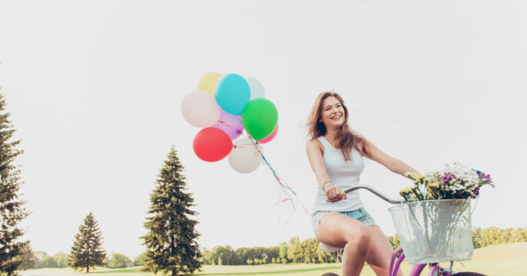 A joyful girl riding a bicycle, smiling, and holding balloons.