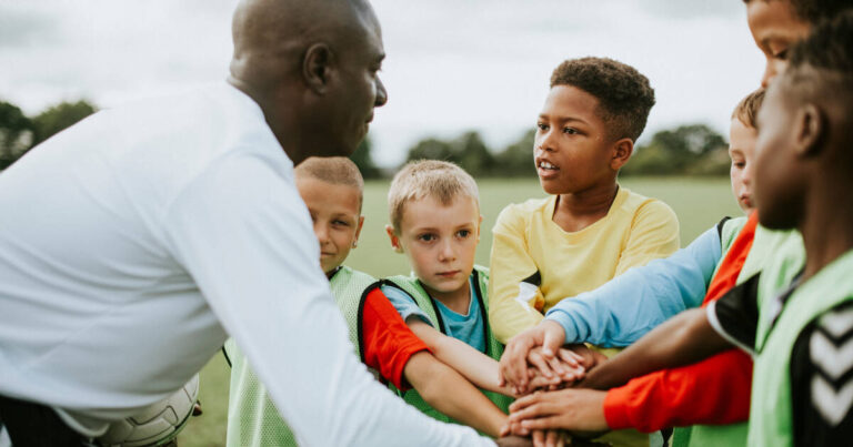 Junior football team forms a huddle, stacking hands before a match, attentively listening to their coach's instructions.