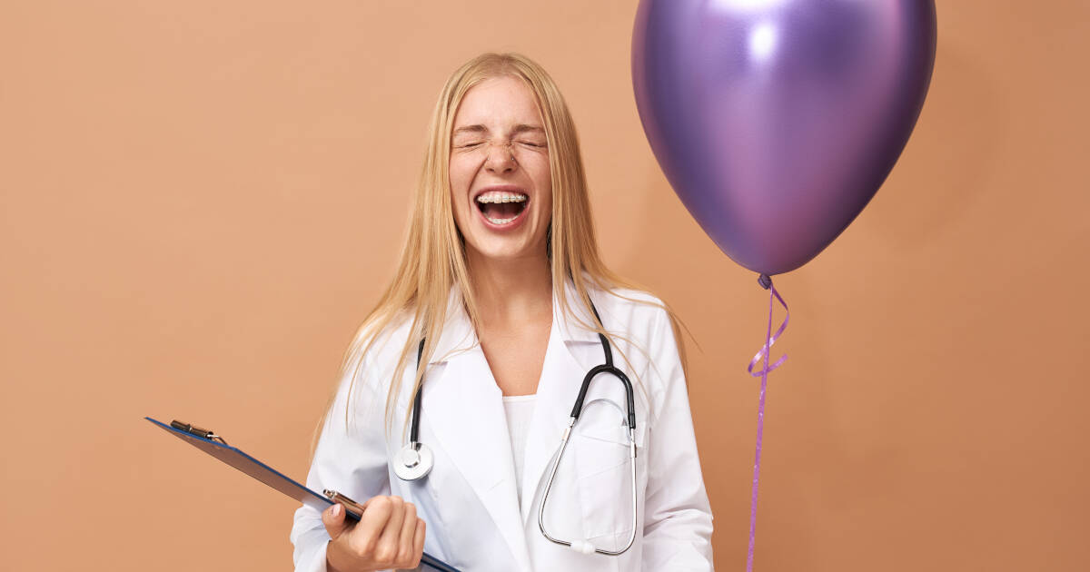 An emotional and joyful young female nurse in a surgical coat laughs with eyes closed as she holds a purple helium balloon.