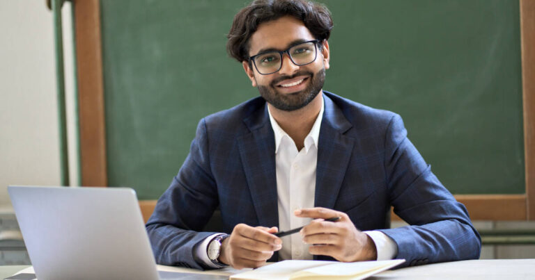Young principal, wearing a suit, sits at desk with laptop, smiling at the camera.