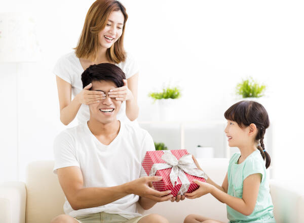 Father surprised by gift from wife and daughter in living room, daughter passes gift box, wife covers eyes, all smiles