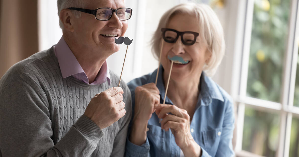 Senior couple sit on couch holding funny party set with carnival accessories, fake eyeglasses, and mustaches on wooden sticks.