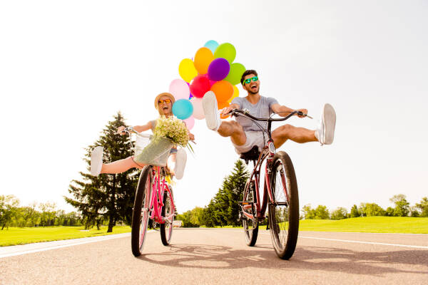 funny young couple rides bicycles in the park, legs raised, with balloons in the background.