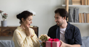 Son surprises his mom with a pink gift box as they sit on the sofa at home