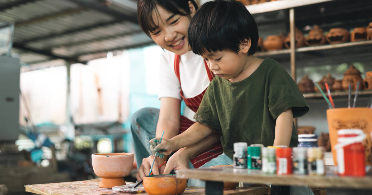 Mother teaches young son how to paint a mug at a ceramics workshop, bonding over creativity