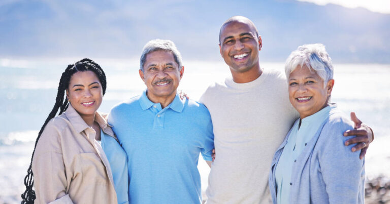Daughter stands next to her dad, her boyfriend next to him, and mom on the end, standing on the beach posing and smiling