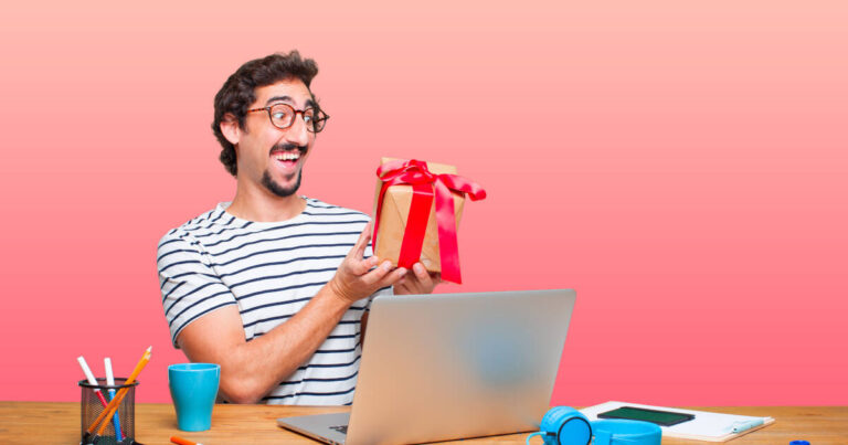 A man with glasses, wearing a striped shirt, sits at a desk with a laptop, markers, and headphones. He is smiling excitedly while holding a wrapped gift with a red ribbon. The background is a gradient from light pink to peach.