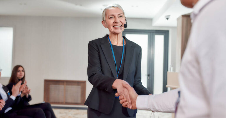 Woman smiling wearing a black suit and a microphone headset, shaking hands with another person wearing a white shirt.