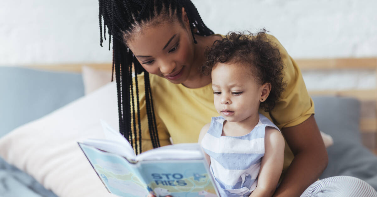 Mother sitting on bed, reading book to adorable baby daughter.