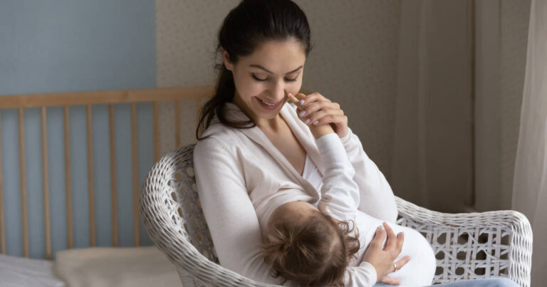 Happy, young Mom enjoying breastfeeding her baby, sitting on a white wicker chair.