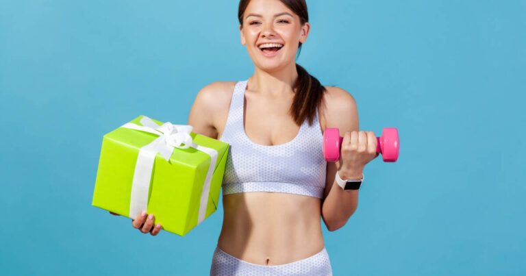 Smiling woman in work out gear holding a green gift box with white ribbon in one hand and a pink dumbbell in the other.