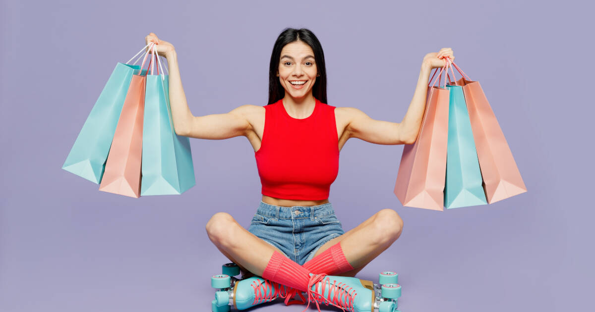 A cheerful woman in a red crop top and denim shorts sits cross-legged on roller skates, holding multiple pastel shopping bags in both hands against a purple background.