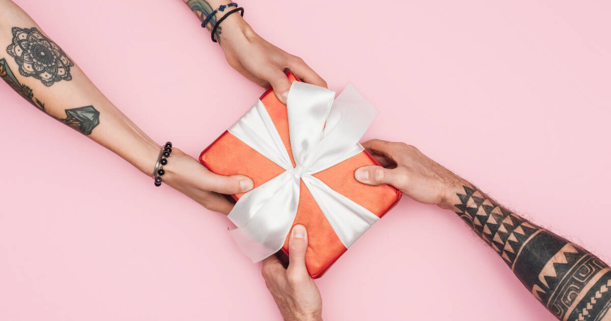 A cropped view of tattooed hands holding a gift box against a pink background.