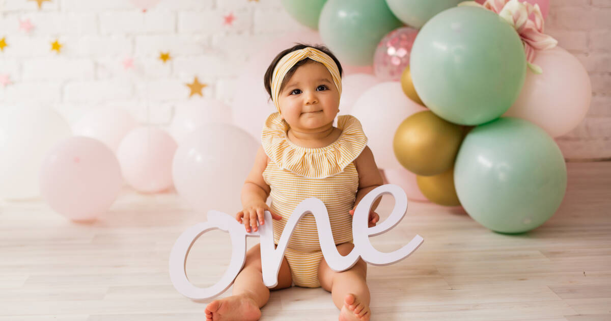 A baby girl sits on a wooden floor, holding wooden lettering that spells 'ONE', with a balloon in the background.