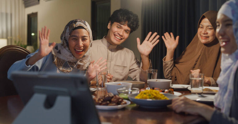 A smiling family sits around a dinner table with plates of food and waves toward a tablet during a video call.