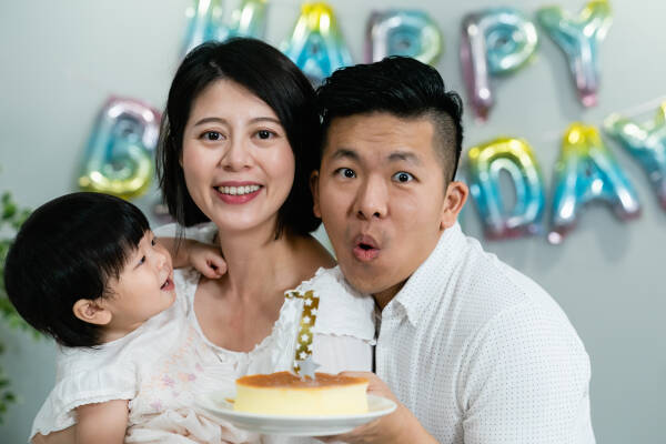 mom holds the child as dad blows out the '1' candle on the cake, with a 'HAPPY BIRTHDAY' balloon in the background