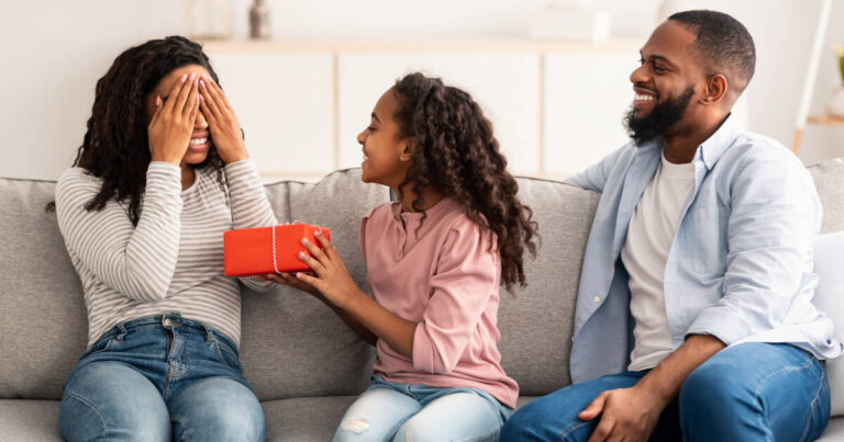 Young girl surprises her mum with a gift on Mother's Day, while dad smiles on the sofa next to them.