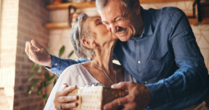 An elderly couple happily exchanges gifts at home, with a surprise and a kiss on the face.