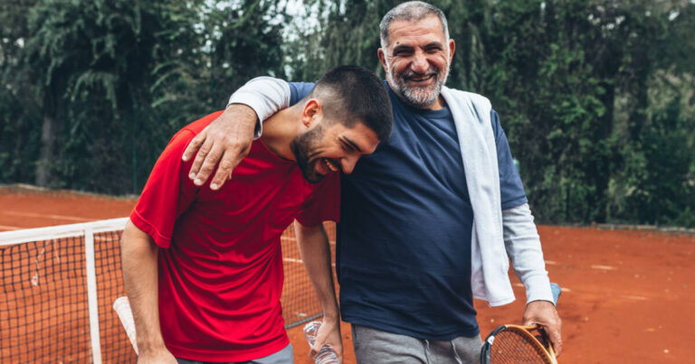 Father and son smiling on a tennis ground, father has his arm wrapped around his son.