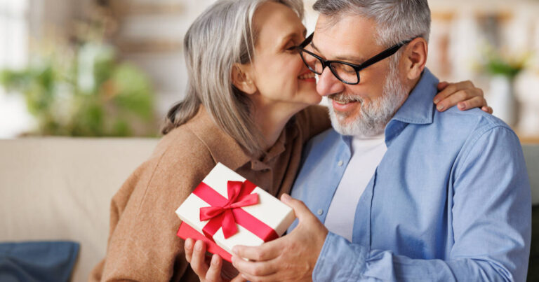 A loving senior woman gives a gift box to her surprised and happy husband. They sit together on the couch at home, with the mature man smiling as he receives the present.