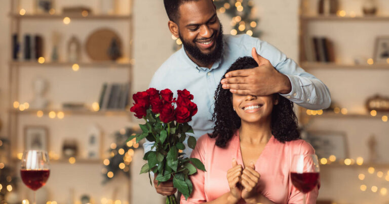 Smiling man covers his woman's eyes, presenting her with a bunch of red rose.