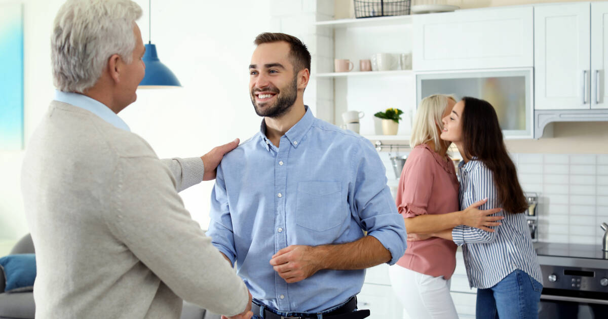 Husband shakes hands with father-in-law while wife hugs her mother in the background, all in the kitchen.