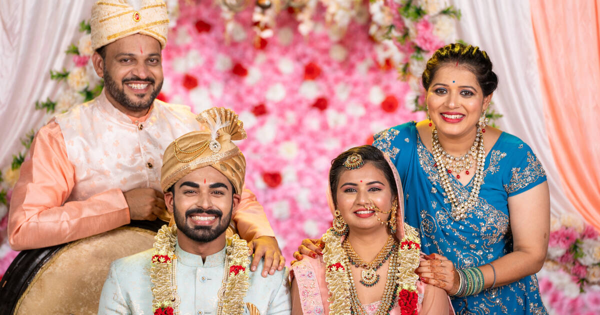 Happy Indian middle-aged parents stand smiling behind the bride and groom at a wedding, celebrating together in a joyful and festive moment.