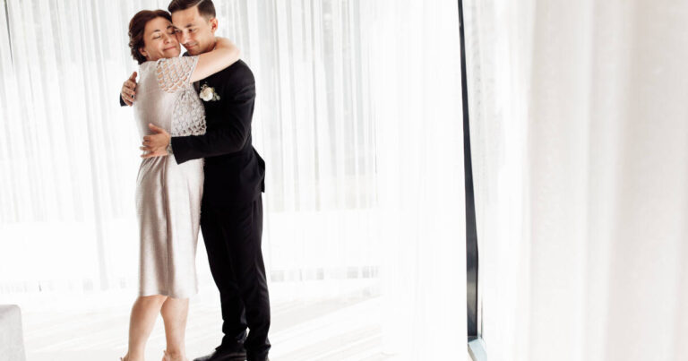 Groom hugs his mother on his wedding day in a white room, both looking happy.