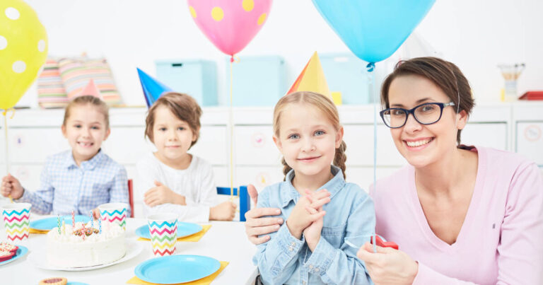 Kindergarten teacher embraces girl in party hat, surrounded by balloons and party food, with two other children nearby