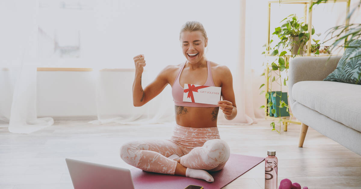 Woman in pink tracksuit does yoga with laptop, holds gift certificate, and celebrates with winner gesture at home