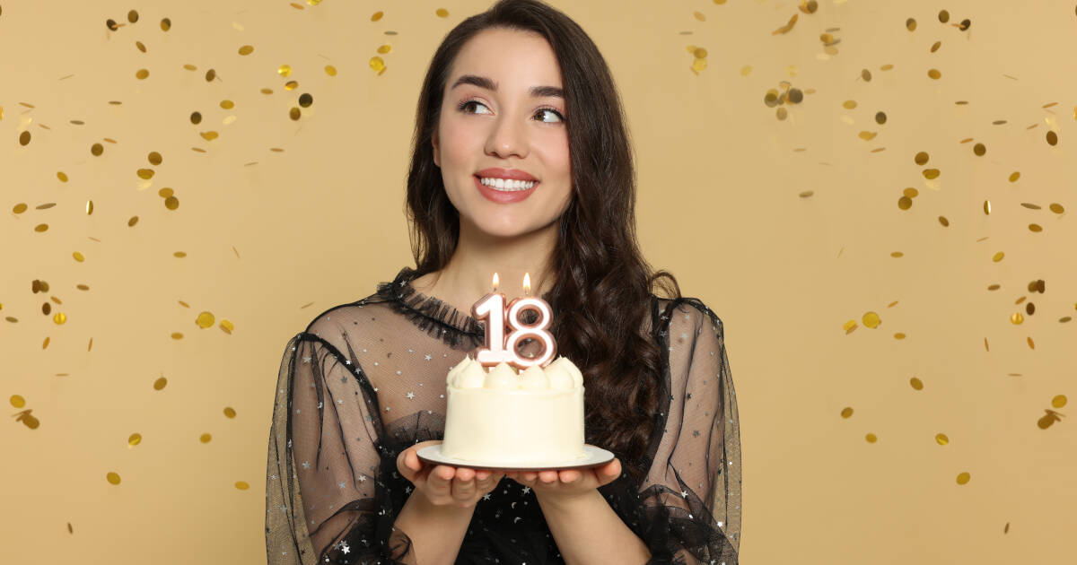 Woman holds a cake with '18' candles as she smiles against a golden backdrop, surrounded by confetti.