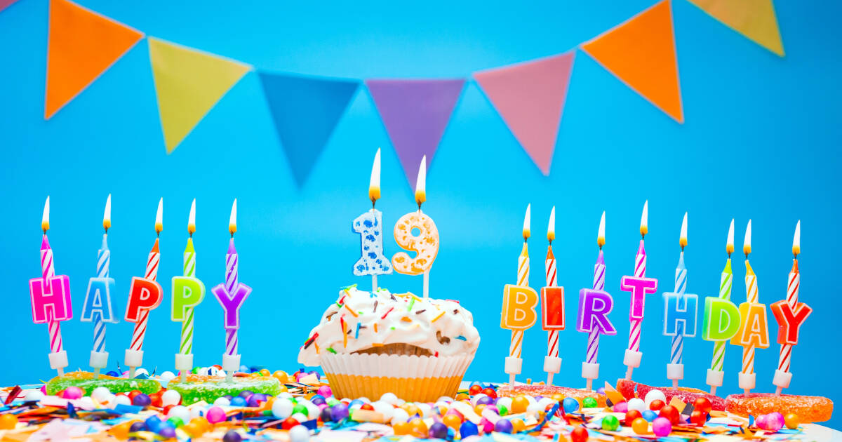 Candles spell out HAPPY BIRTHDAY on a table next to a cake with '19' candles against a blue background.