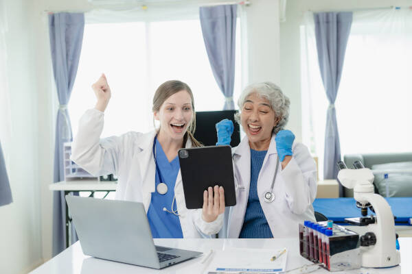 2 successful female doctors celebrates a successful treatment in the hospital, sitting at their desk with arms raised and cheerful smiles.