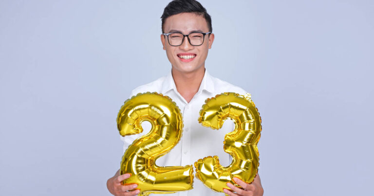 The 23-year-old man smiles as he holds golden '23' balloons against a bluish-gray background.