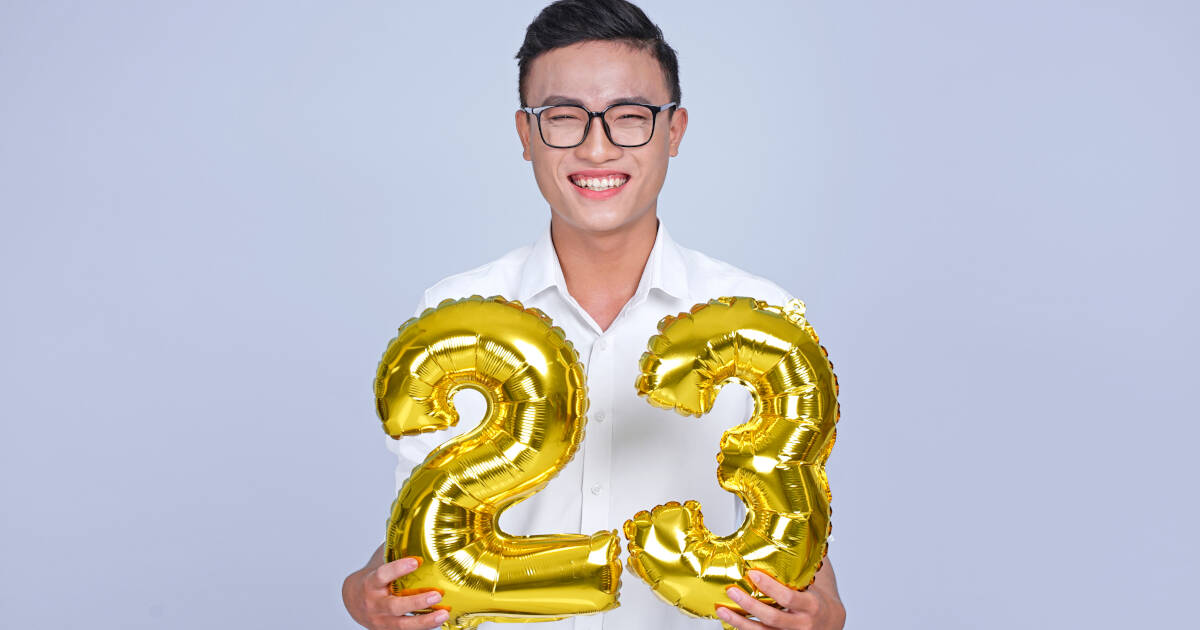 The 23-year-old man smiles as he holds golden '23' balloons against a bluish-gray background.
