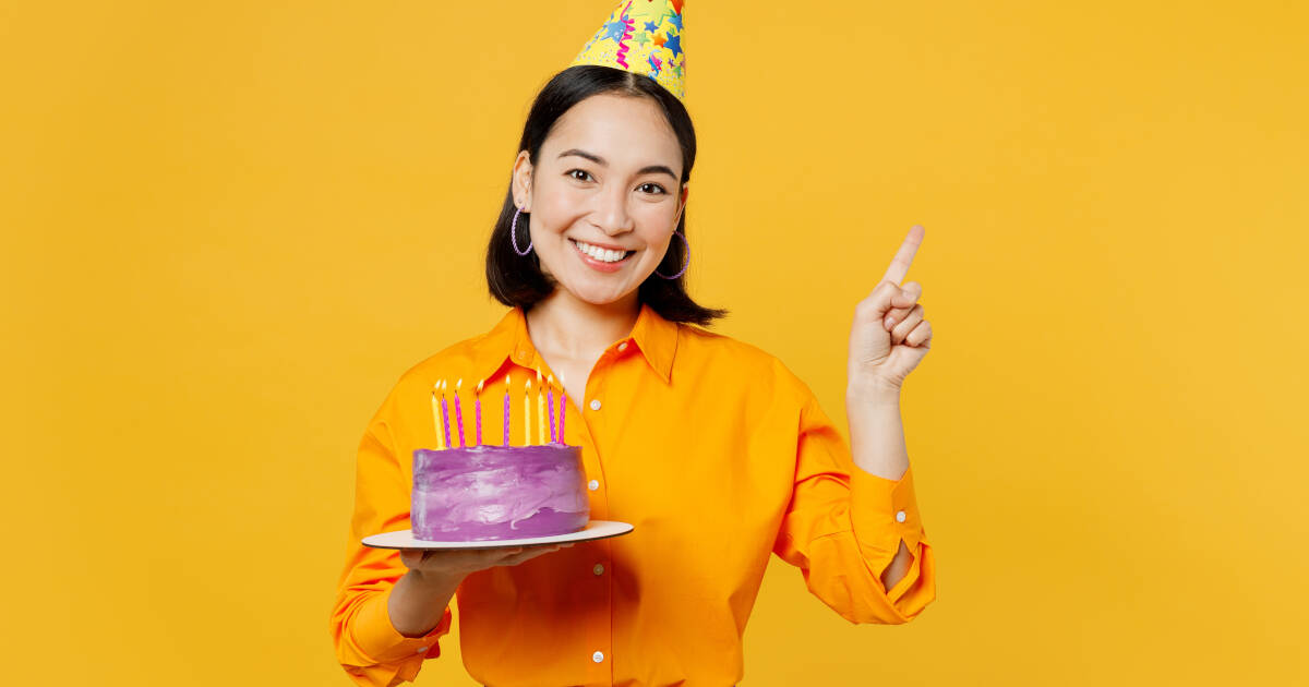 Young woman smiles and points upwards while holding a birthday cake and wearing a party hat against a yellow backdrop.