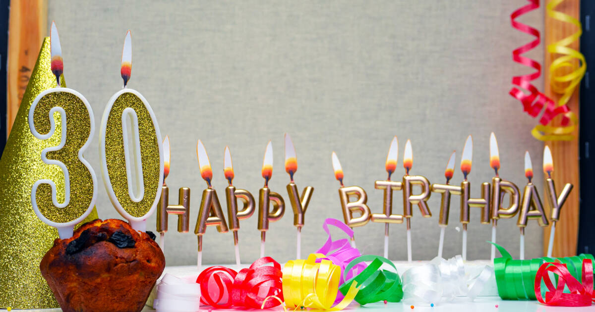 A '30' candle in a cupcake next to HAPPY BIRTHDAY candles on a white table, with decorations in the background.