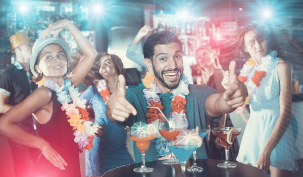 31-year-old man points to cocktails on the table at a Hawaiian-themed party, with guests in the background.