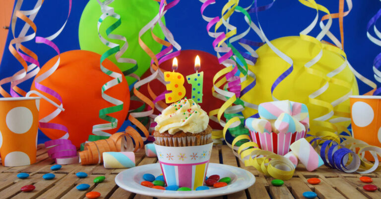 Birthday cupcake with '31' burning candles on a rustic wooden table, surrounded by colorful balloons, plastic cups, and candies.