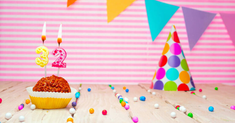 Cupcake with '32' candles lit next to a party hat against a striped pink background, with party bunting against the wall.