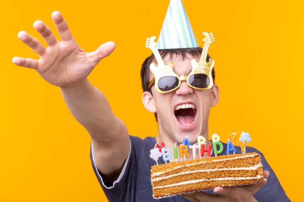 A 19-year-old wearing guitar-shaped glasses and a party hat, holding a birthday cake and pulling a funny expression.