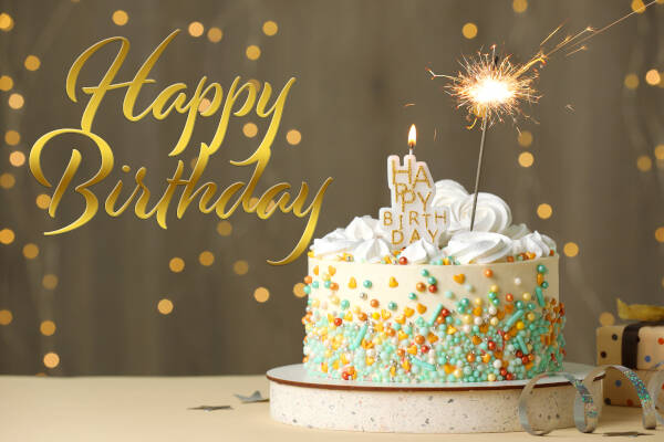 A beautiful birthday cake with a sparkler on it, with writing saying 'HAPPY BIRTHDAY' beside it.