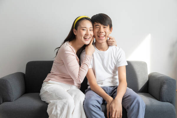 A brother and sister sit on a sofa at home. The sister smiles and holds her brother's cheeks, gesturing for him to smile too.