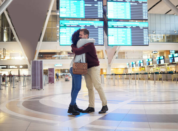 A couple embraces in an airport terminal, with flight information screens visible in the background. The man wears a maroon sweater and the woman has curly hair and a brown purse.