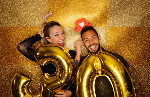 A couple holding golden '30' balloons and smiling widely against a sparkly gold backdrop.