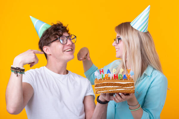 A couple stands with a birthday cake, smiling and pointing against a yellow background.